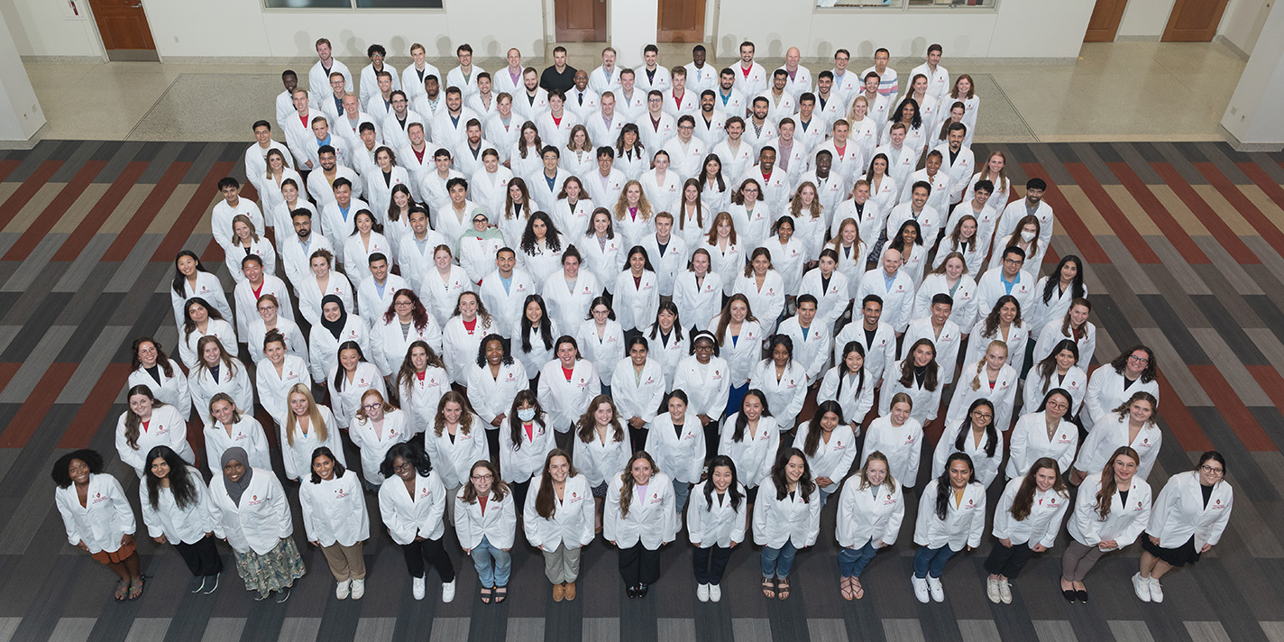 The class of medical students who matriculated in August 2024, wearing their white coats, and looking upward to the photographer in the Health Sciences Learning Center Atrium.